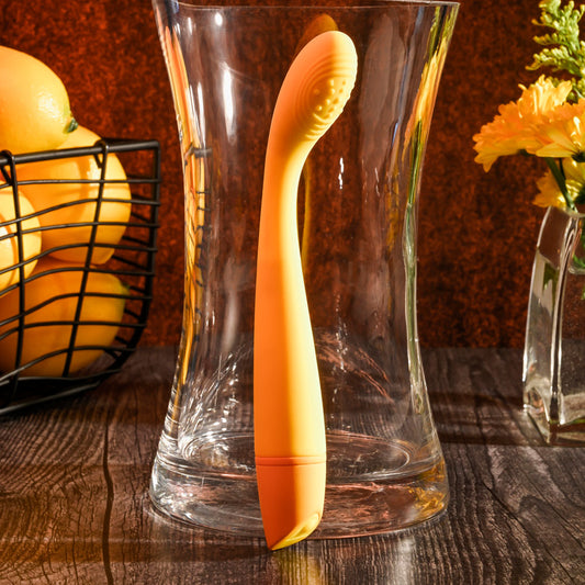 Orange spoon leaning against a clear glass vase on a wooden surface with fruits and flowers in the background.