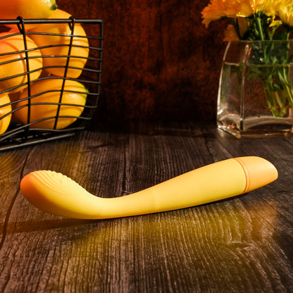 Yellow object resembling a banana on a wooden surface with fruits and flowers in the background.